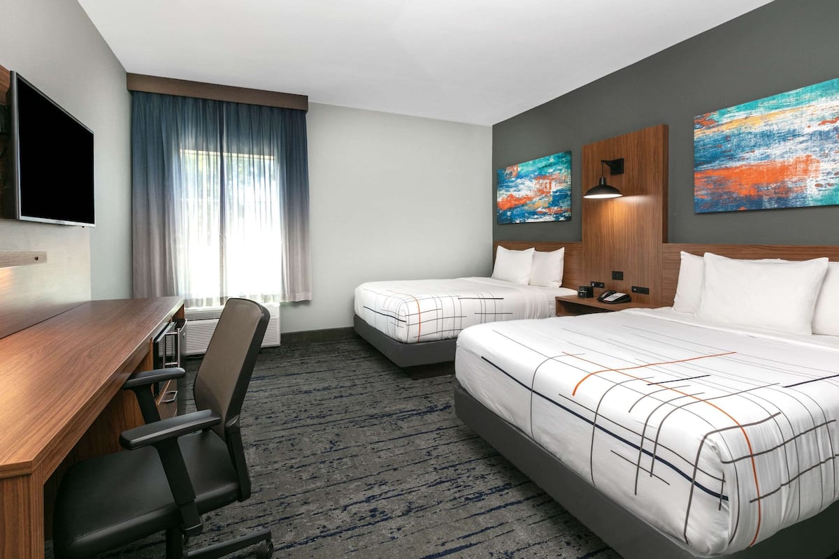 A hotel room with a desk, two beds, a TV, artwork, and a window with curtains.