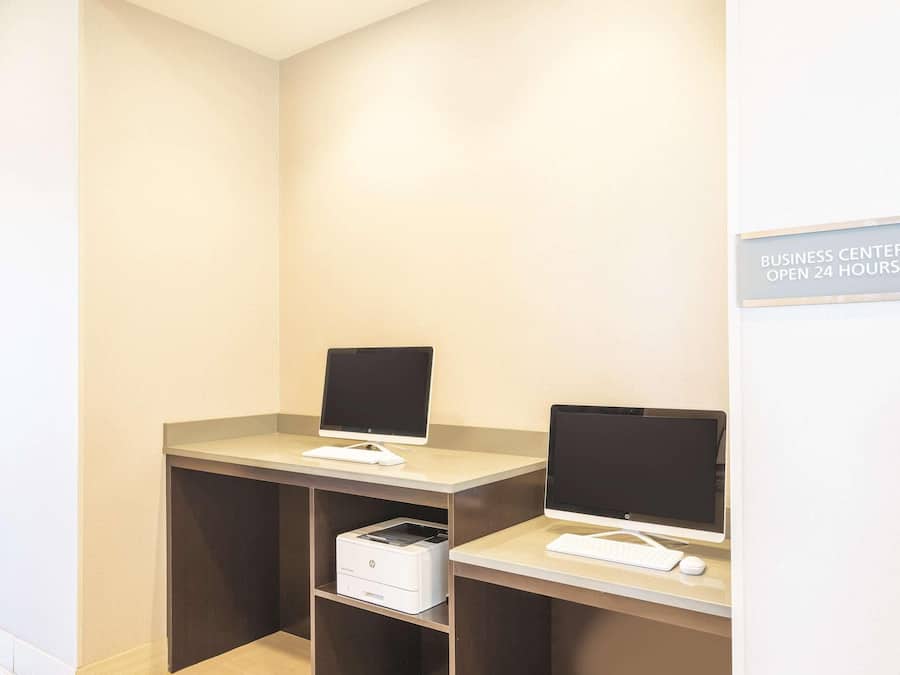 Ein Business Center mit zwei Arbeitsplätzen, jeweils ausgestattet mit einem Bildschirm, einer Tastatur und einer Maus. Ein Drucker steht auf dem Boden. Ein Schild weist darauf hin, dass das Business Center rund um die Uhr geöffnet ist.