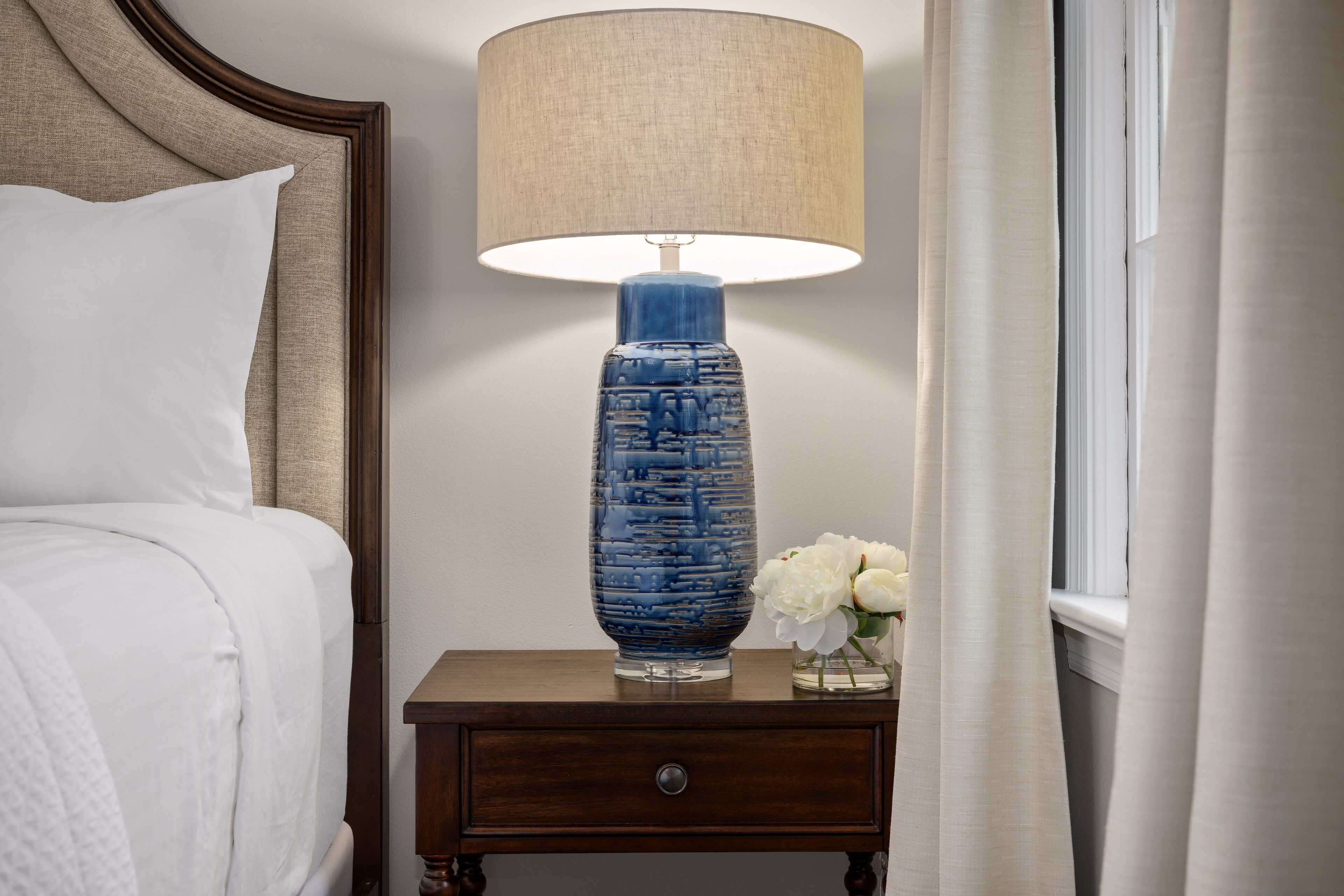A neatly made bed with white linens, a wooden nightstand with a lamp, and a vase with white flowers.