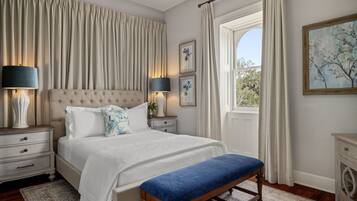 A bedroom with a tufted headboard, a large bed with white linens, a blue bench, two bedside tables with lamps, a window with curtains, and framed artwork on the wall.