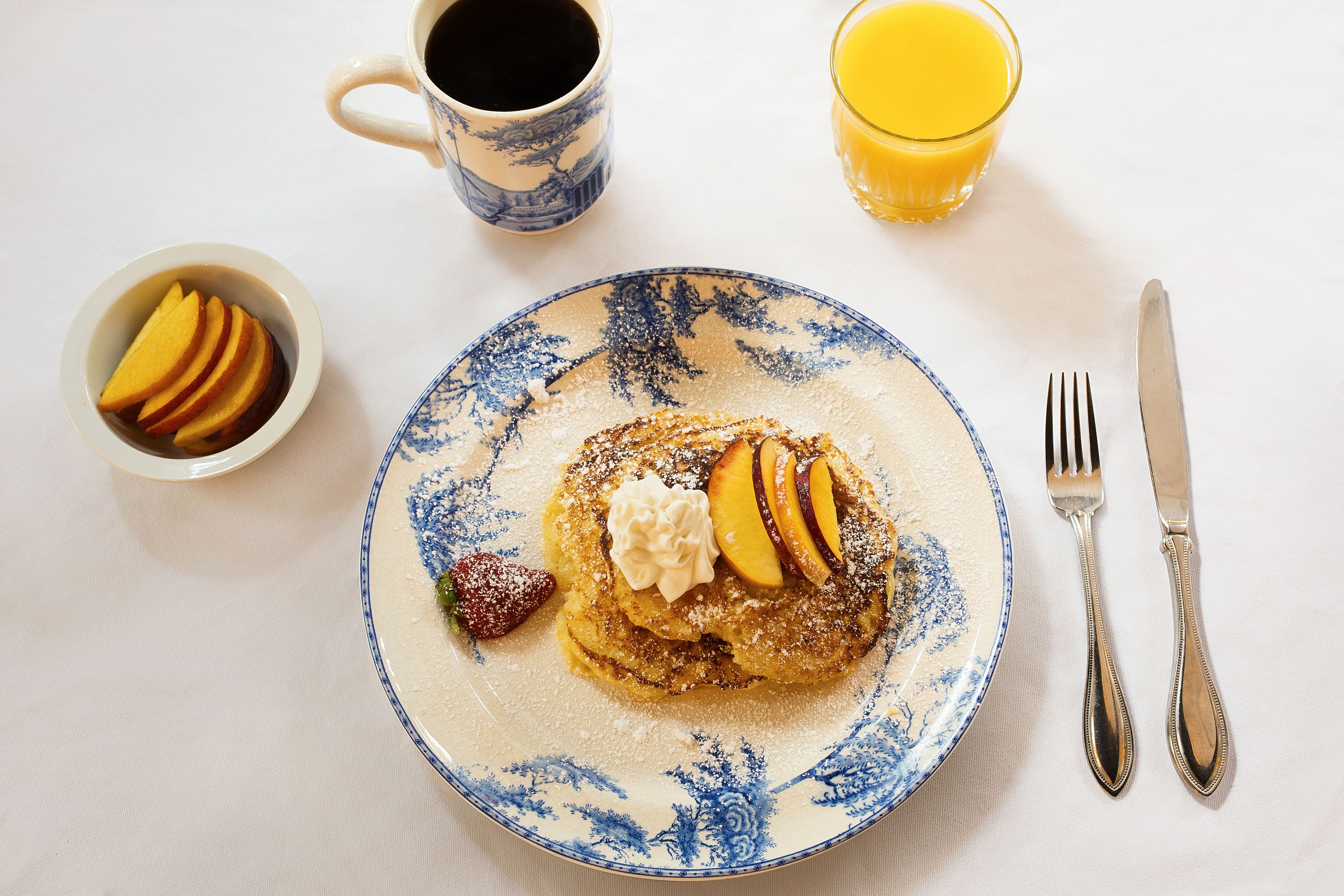 A breakfast plate with pancakes, fruit, and whipped cream, accompanied by a coffee cup, a glass of orange juice, and a bowl of pancakes.