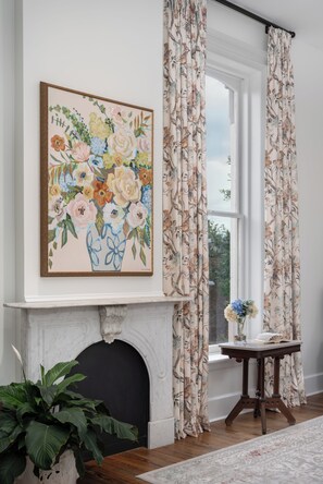 Una habitación con chimenea, cortinas florales, un cuadro con flores enmarco y una mesita con un florero.