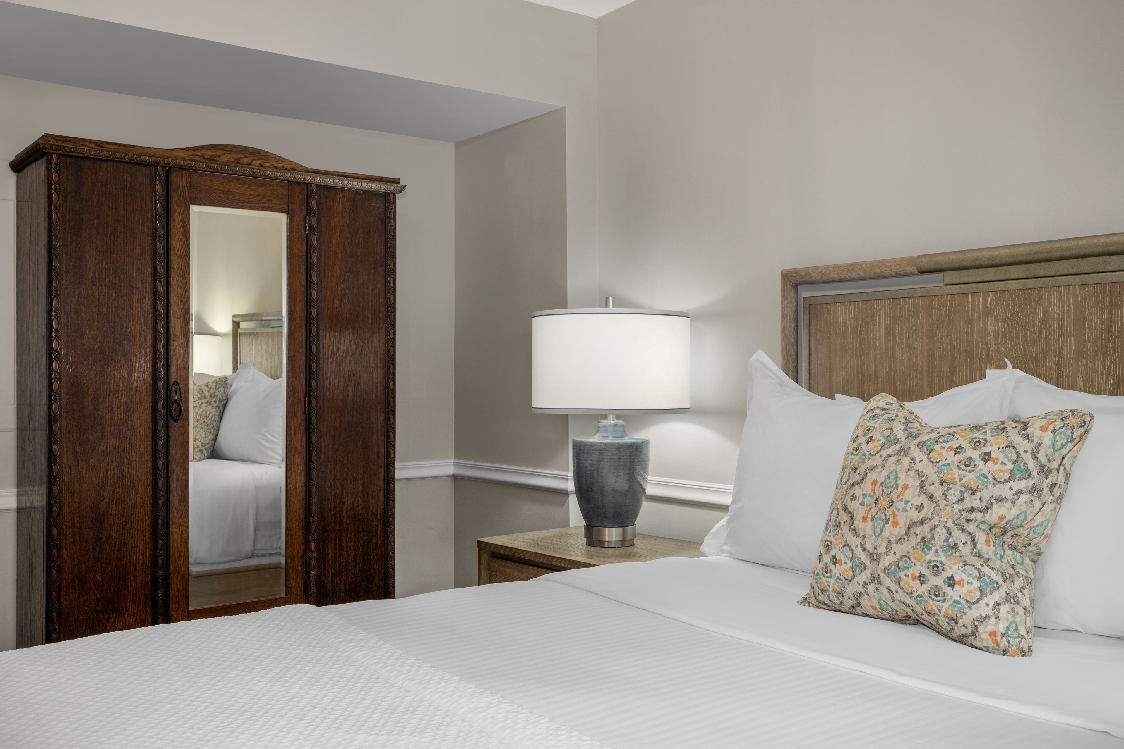 A hotel room with a large bed, a wooden wardrobe, a nightstand with a lamp, and a mirror.