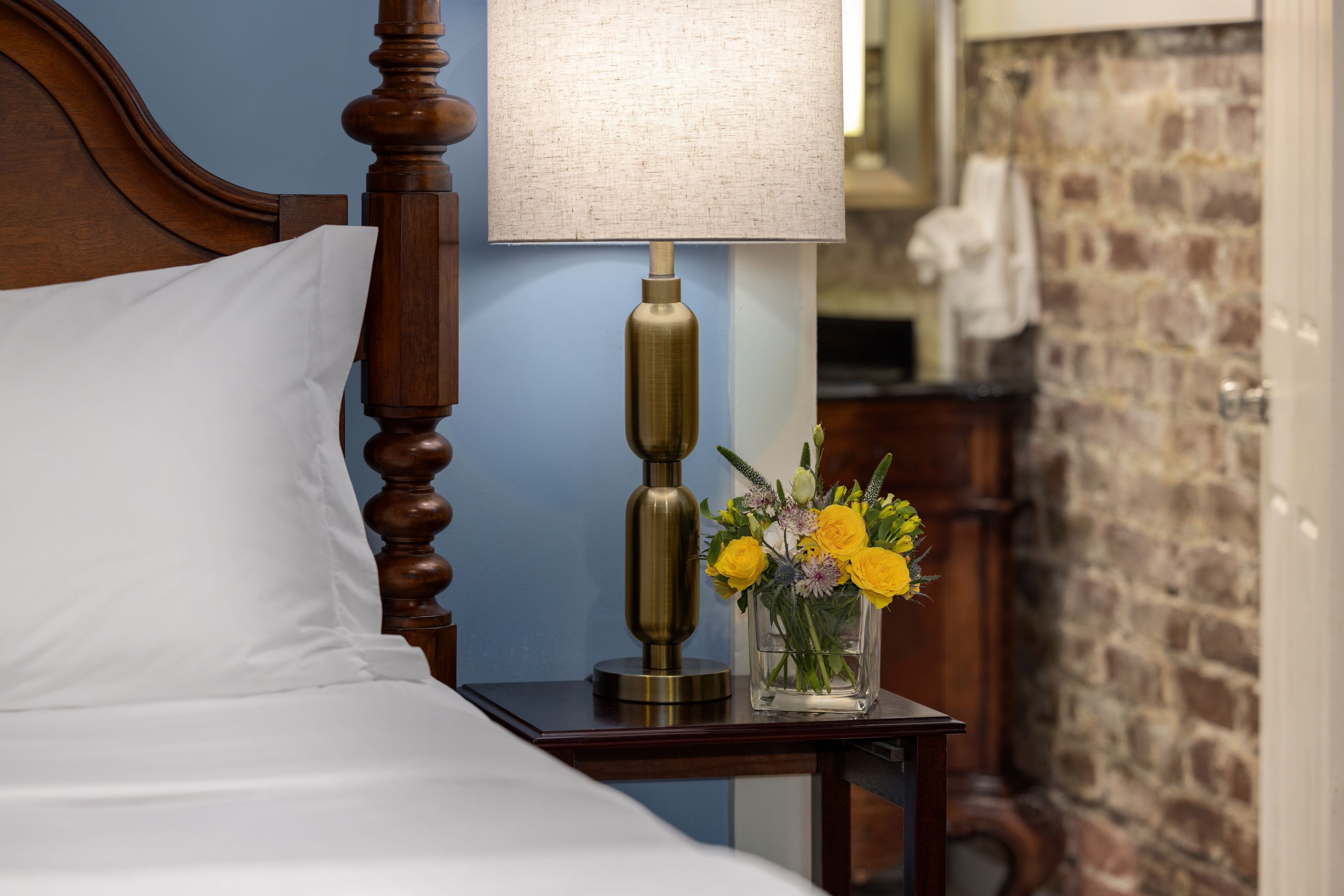 A neatly made bed with a white pillow and a wooden headboard. A brass lamp with a white shade and a vase of yellow flowers are on a wooden nightstand beside the bed.