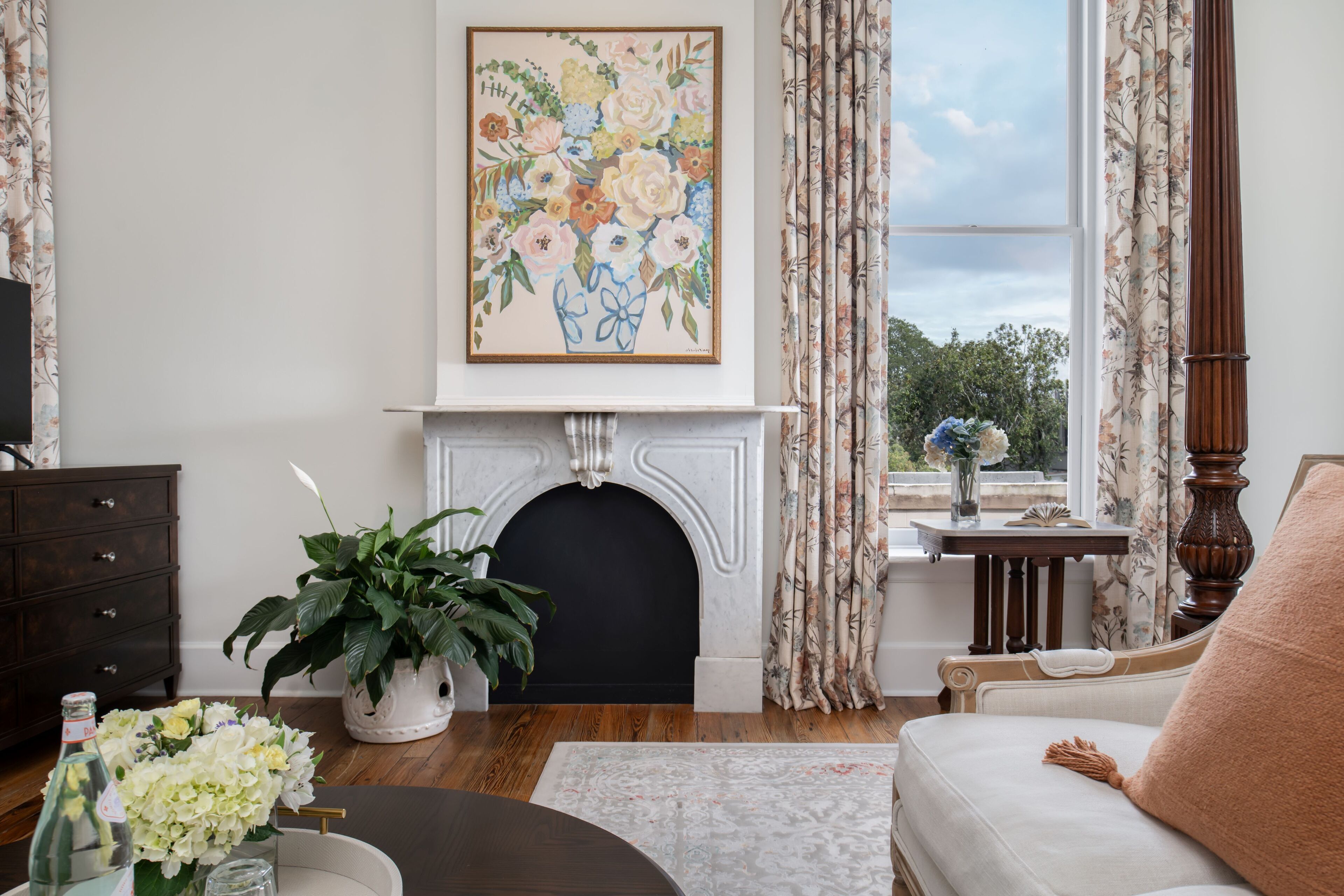 A living room with a fireplace, floral artwork, a sofa, and a view of the outdoors.
