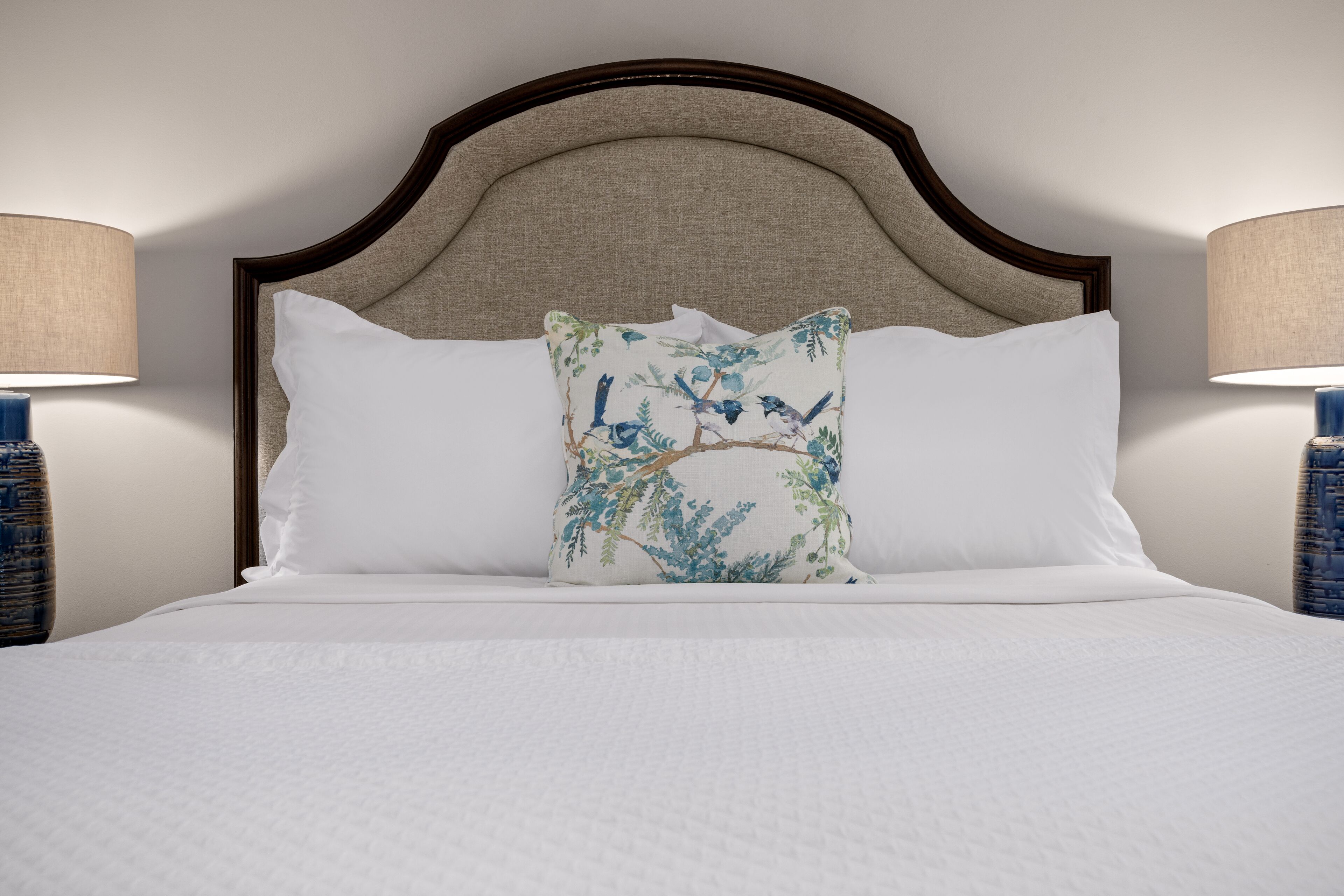 A neatly made bed with white linens, a decorative pillow with a nature-themed design, and a wooden headboard.