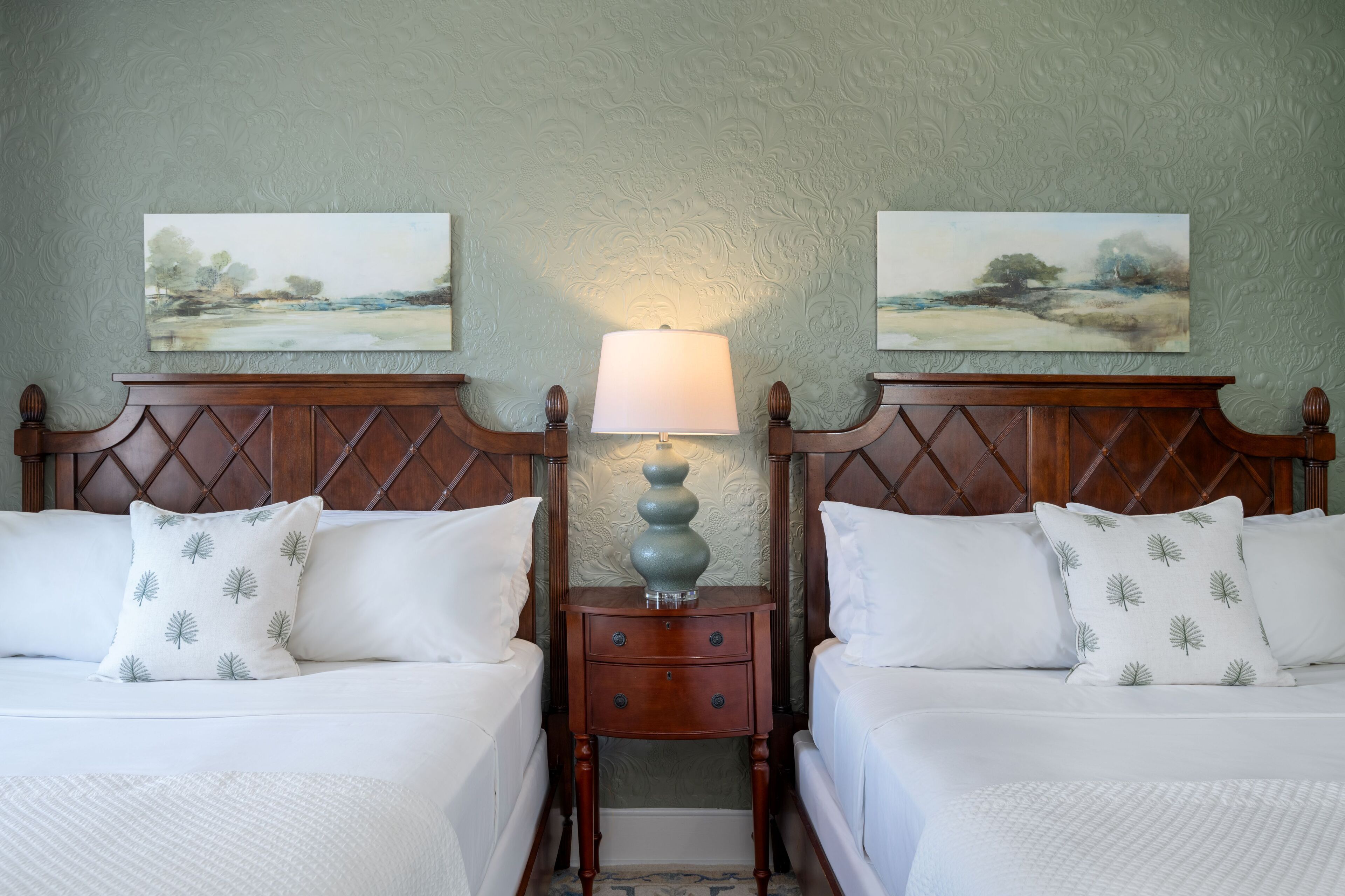 Two beds with white bedding and leaf-patterned pillows, a wooden nightstand with a lamp, and two landscape paintings on the wall.