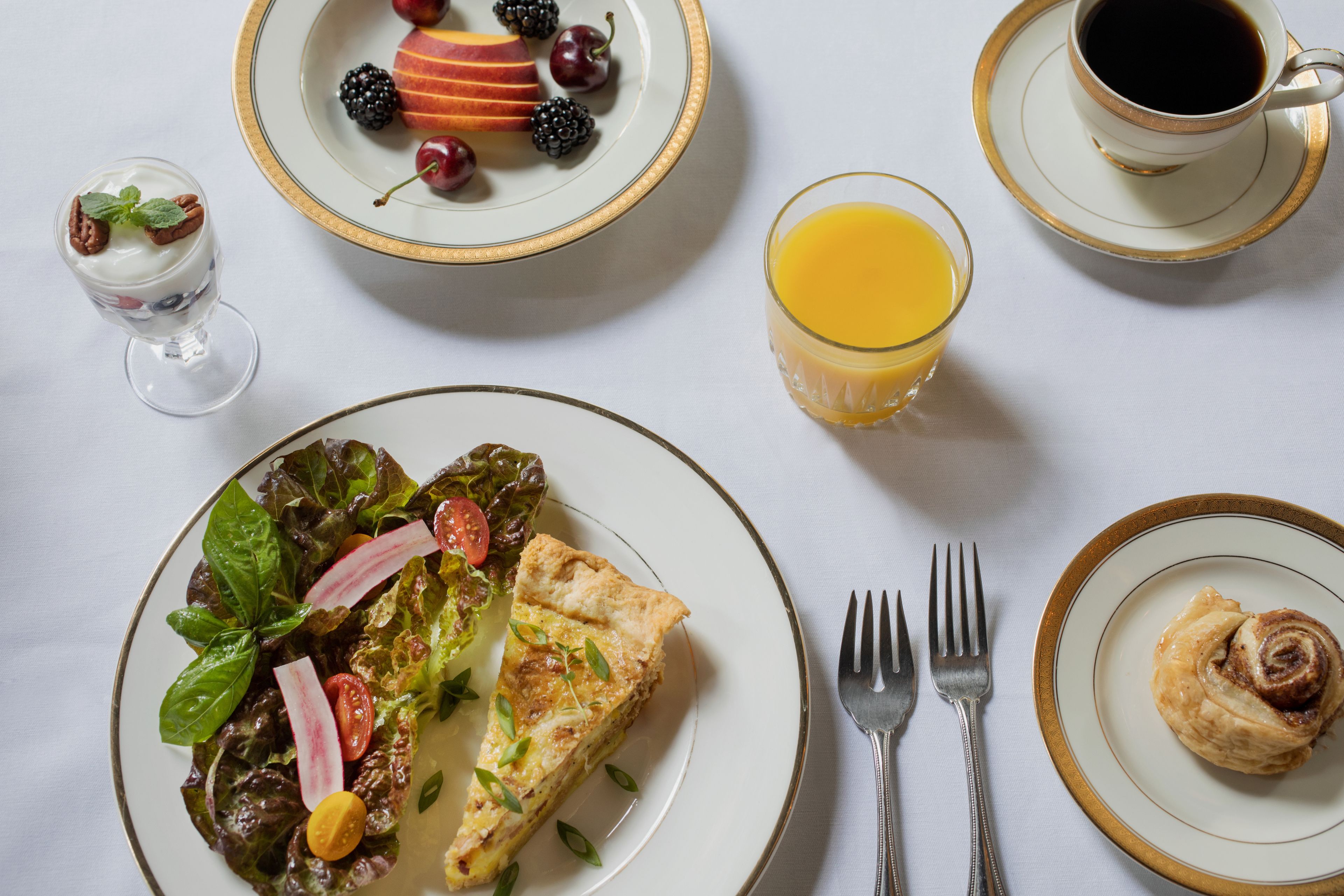 A breakfast setting with a plate of salad, a slice of quiche, a glass of orange juice, a cup of coffee, and a pastry.