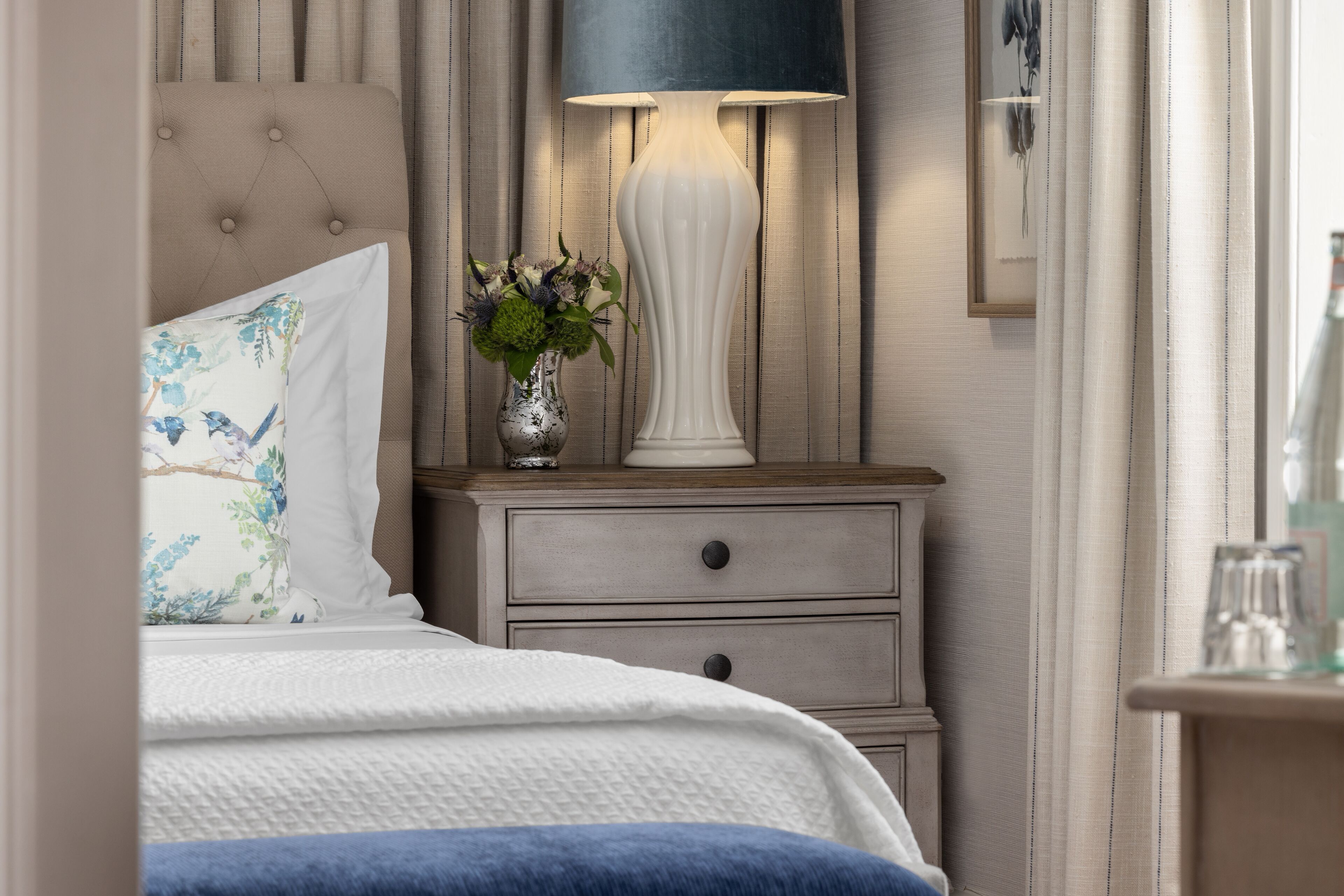 A bedroom with a bed, a nightstand with a lamp, and a vase with flowers.
