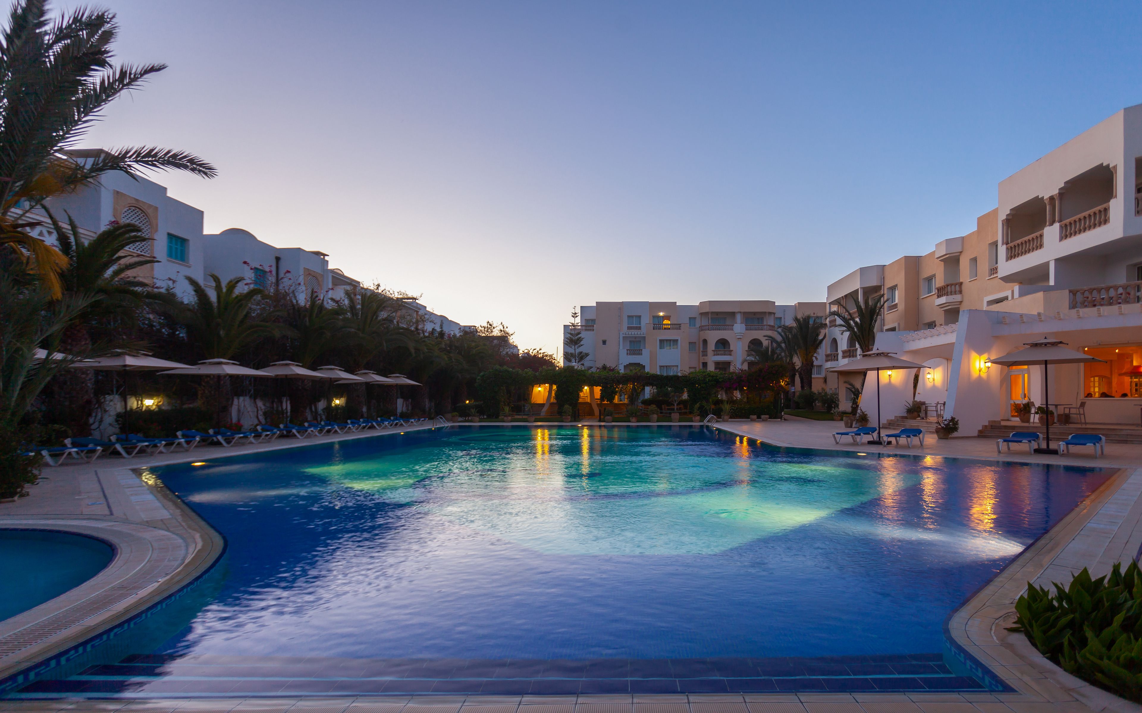Una piscina con sillas de descanso y sombrillas, rodeada de palmeras y edificios con balcones.