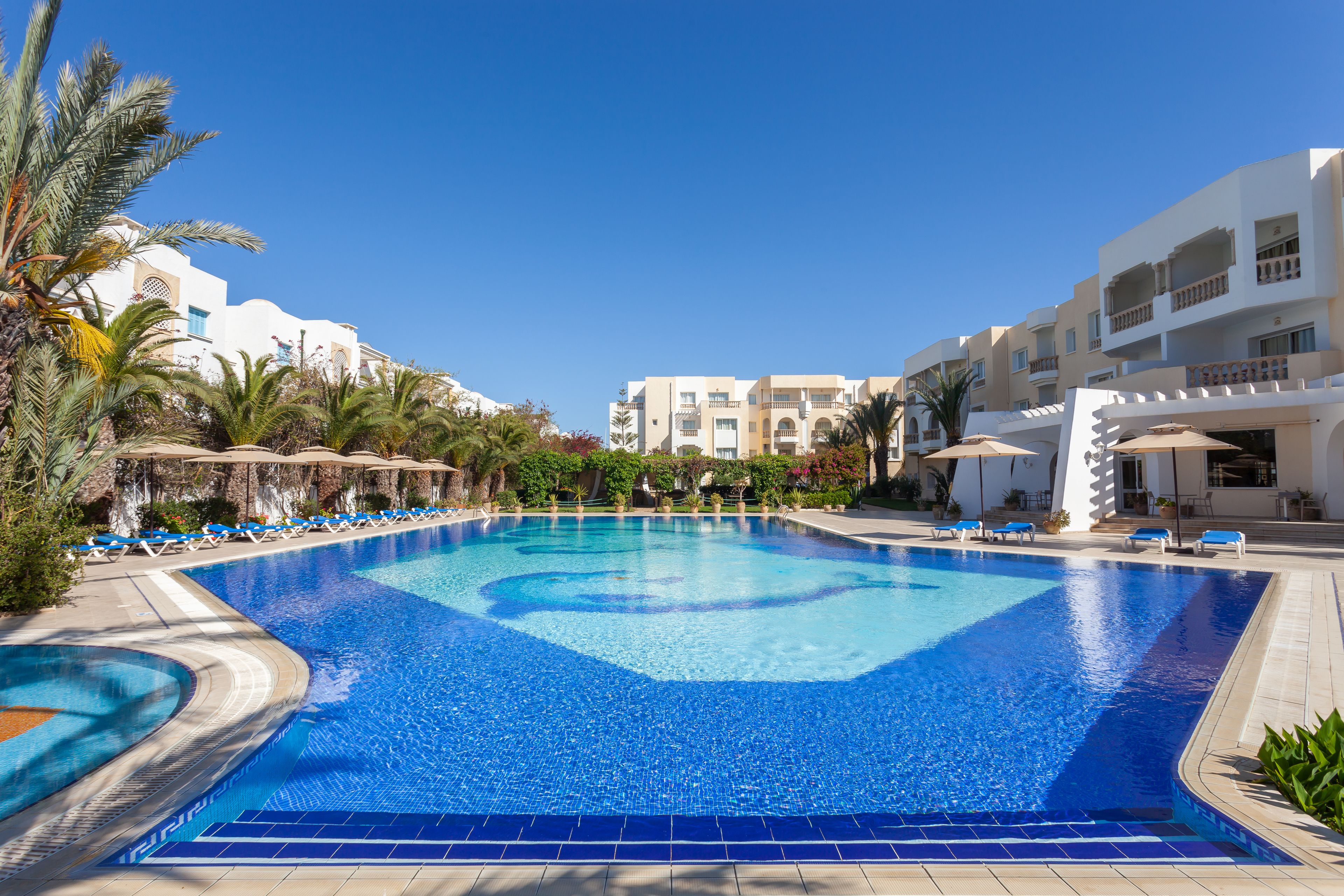 Una piscina grande con baldosas azules, rodeada de sillas de descanso y palmeras.