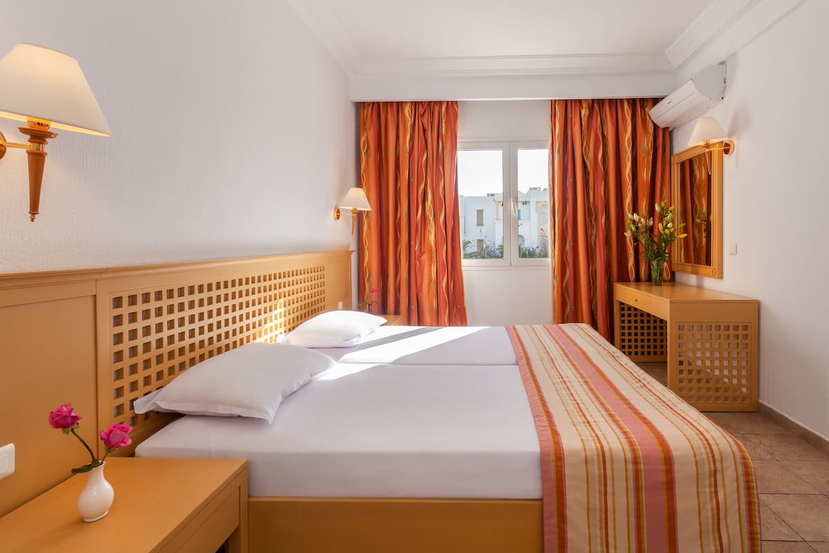 A hotel room with a double bed, a wooden headboard, a nightstand with a vase of flowers, a desk with a mirror, and a window with curtains.