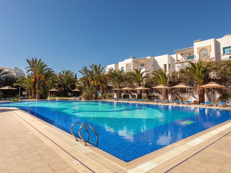 A swimming pool with lounge chairs and umbrellas, surrounded by palm trees and modern buildings.