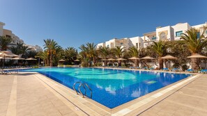 A swimming pool with lounge chairs and umbrellas, surrounded by palm trees and modern buildings. - Corail Apart’hotel & Splash park (Hammamet)