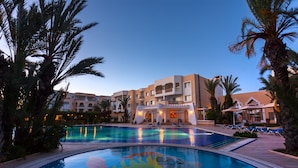 A swimming pool with a palm tree-lined deck, a building with balconies, and a clear sky.