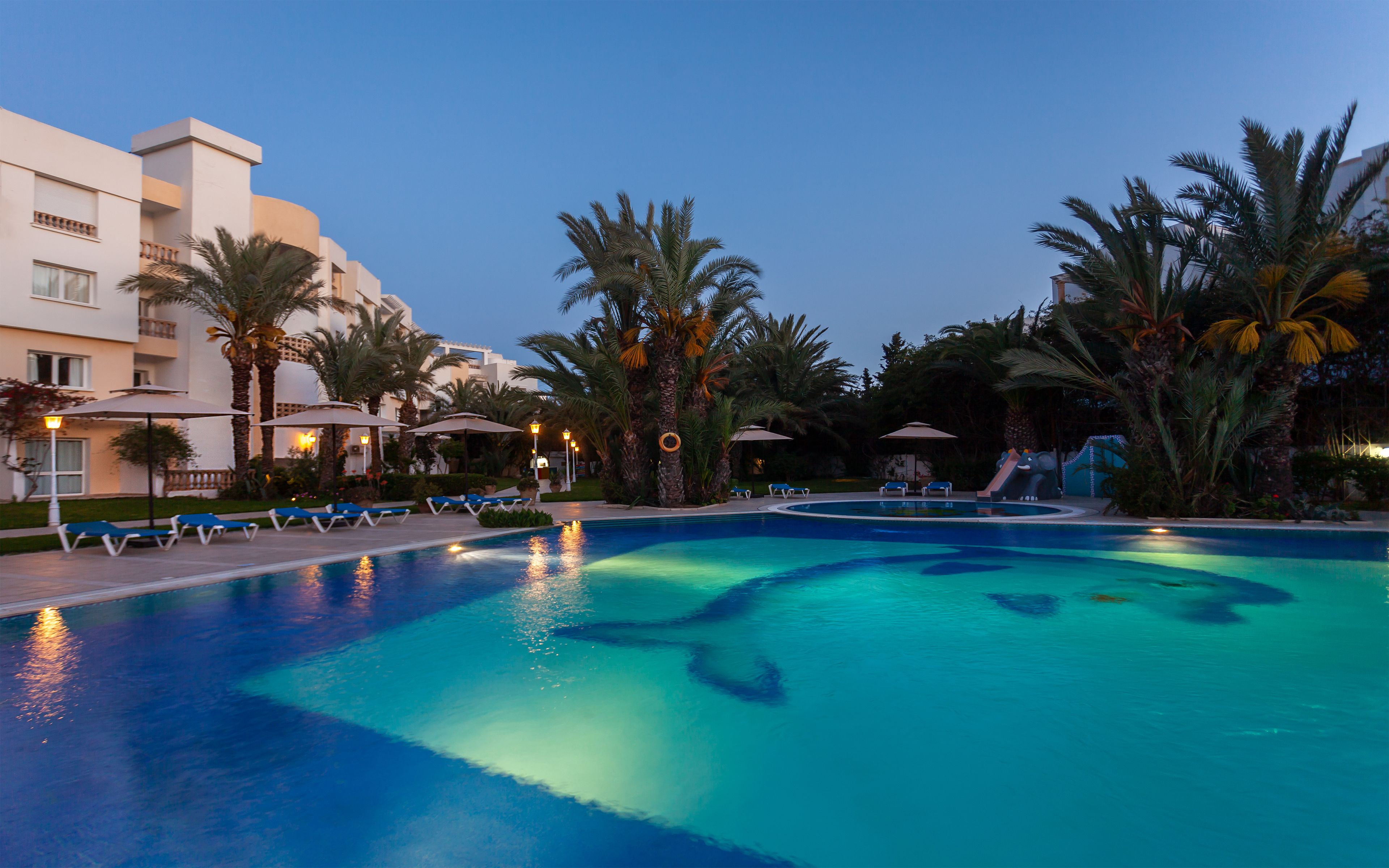 A swimming pool with lounge chairs and umbrellas, surrounded by palm trees and a building with balconies.