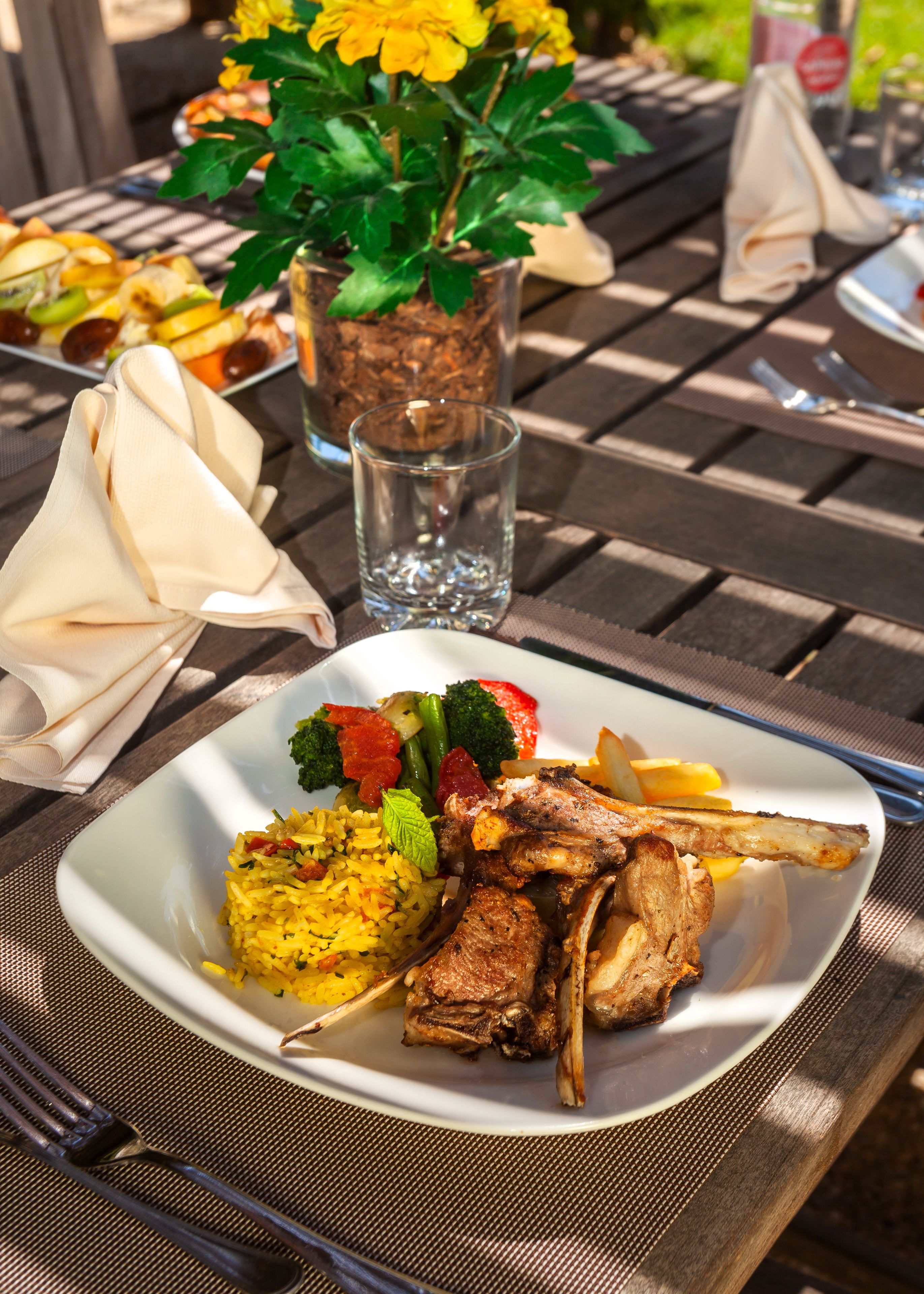A square plate of food with meat, vegetables, and rice, placed on a wooden table with a napkin and a glass of water.