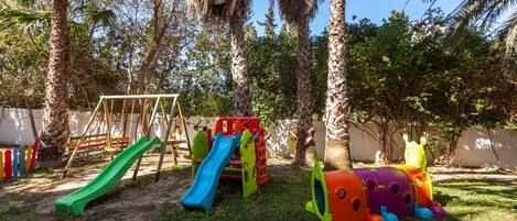 A playground with slides and climbing structures, surrounded by trees and a white wall.