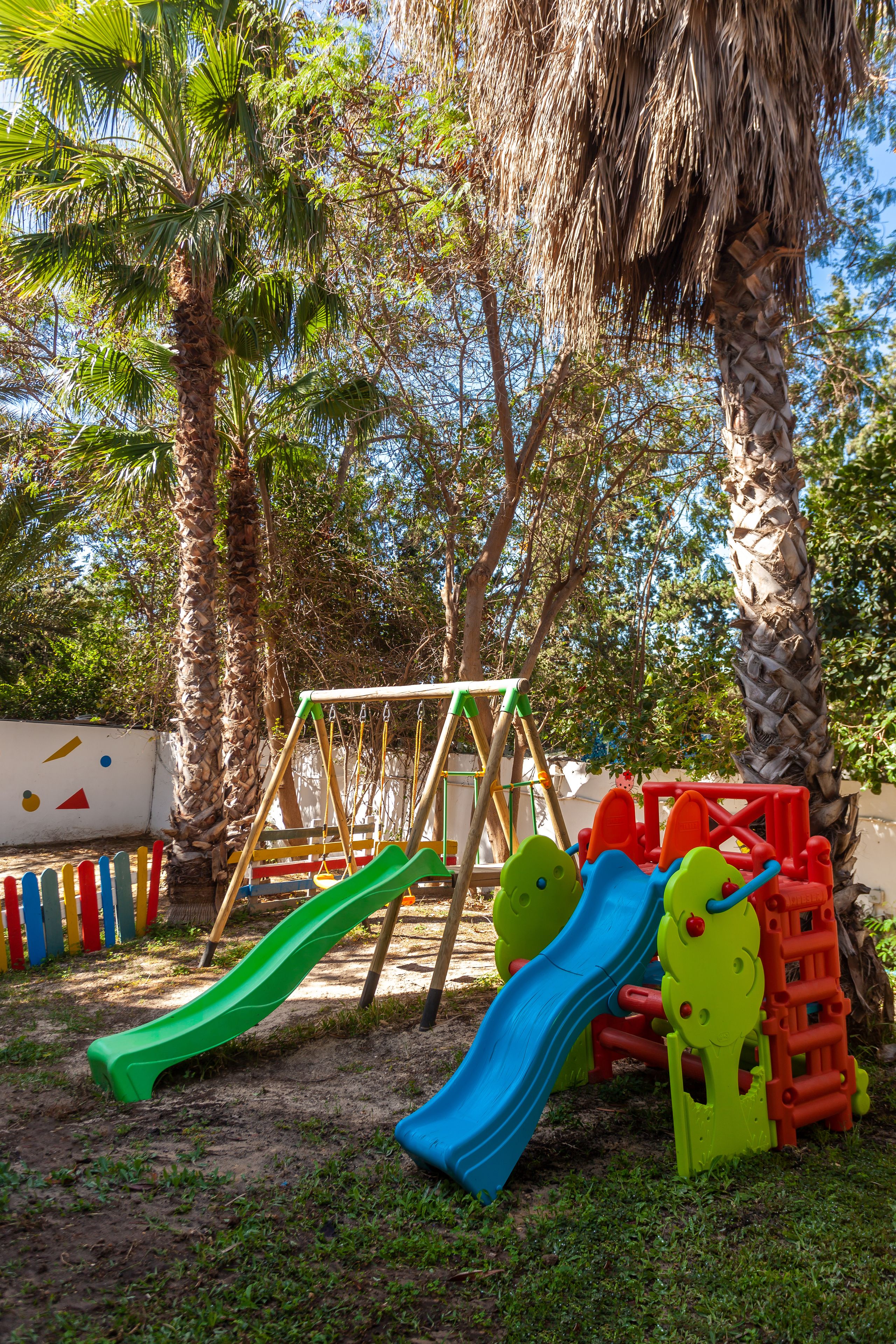 A playground with slides and climbing structures, surrounded by palm trees and a white wall with colorful decorations.