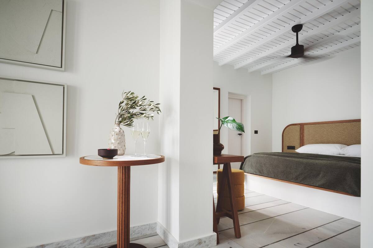 A bedroom with a bed, a bedside table with a vase and glasses, a wooden chair, and a ceiling fan.