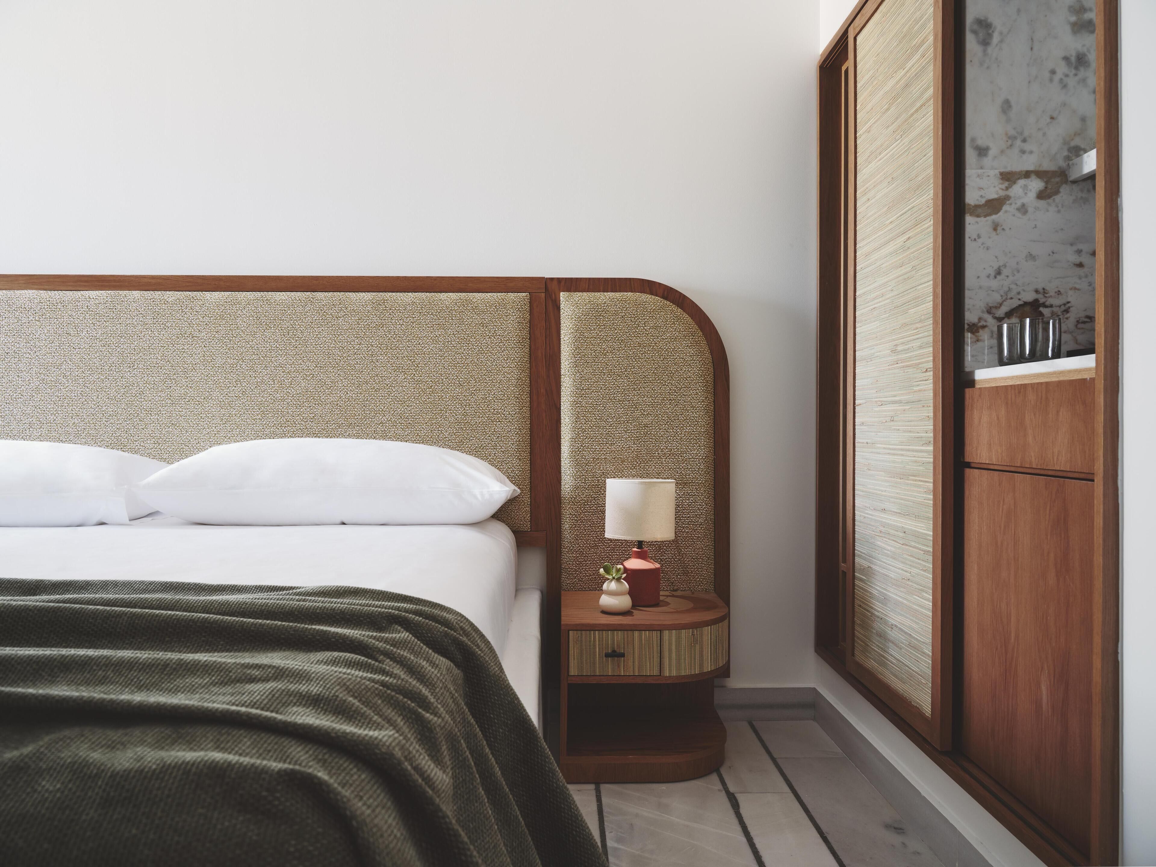 A neatly made bed with white pillows and a green blanket, a bedside table with a lamp and a small decorative item, and a wooden wardrobe with a mirror.