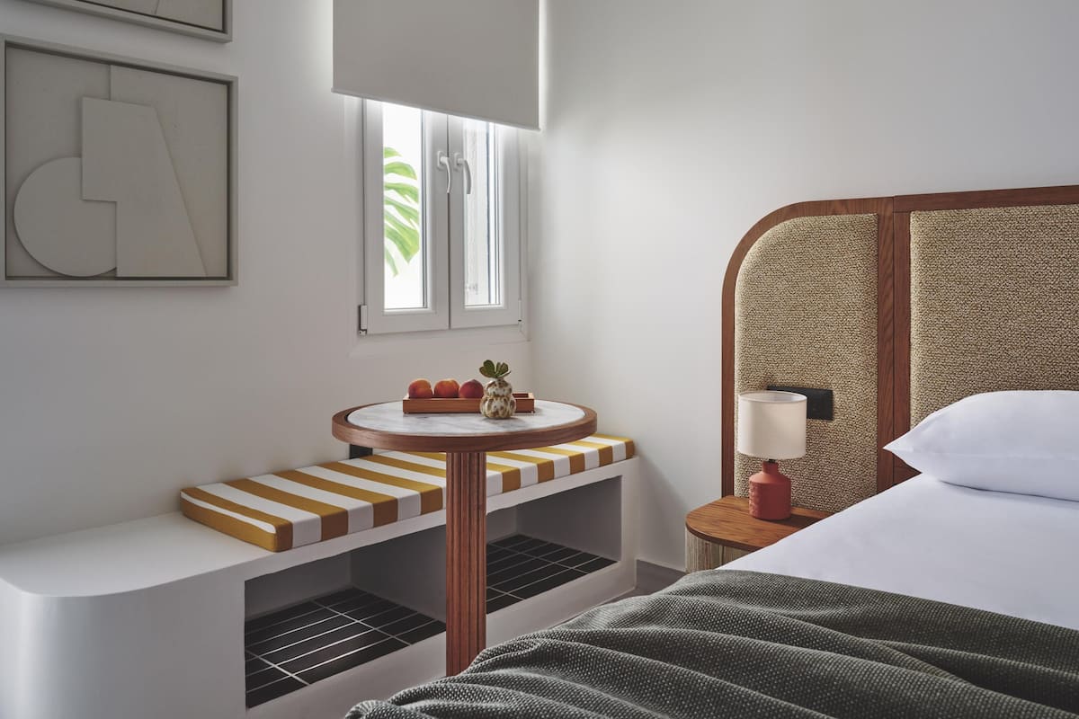 A hotel room with a bed, a bedside table with a lamp, a small round table with fruit, a bench with striped cushions, and a window with a view of greenery.