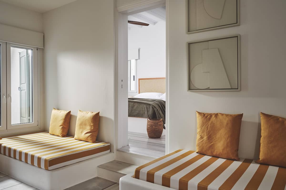 A modern hotel room with a large window, a built-in bench with cushions, a bed with a headboard, and a small table.