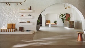 Une pièce spacieuse avec un plafond haut, des murs blancs et une grande porte en arc qui mène à une autre pièce. On y trouve des étagères blanches où sont disposés divers objets, un coin salon avec des coussins et une petite table sur laquelle est posée une plante.