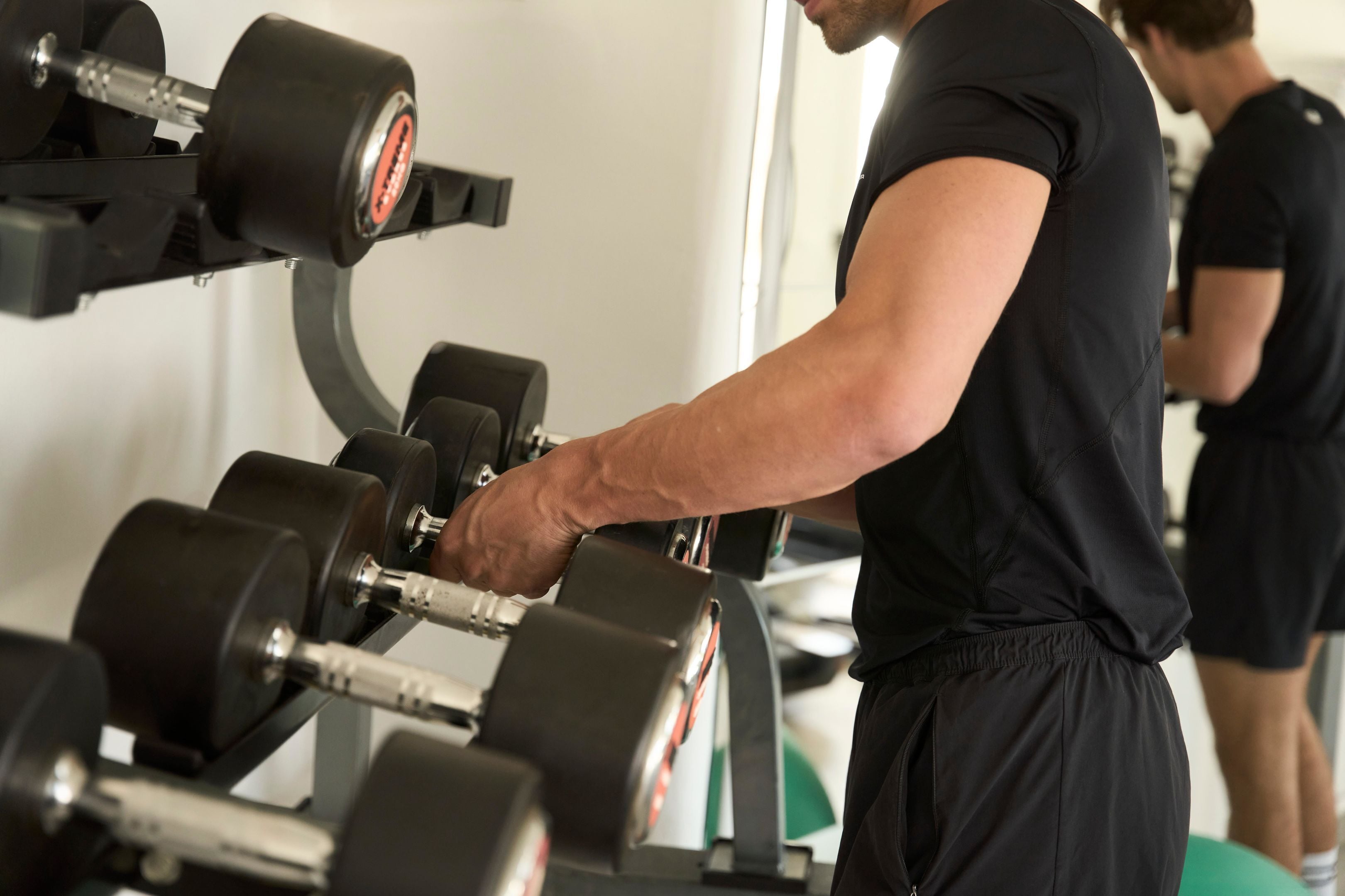 A gym with weightlifting equipment, a person exercising, and another individual in the background.