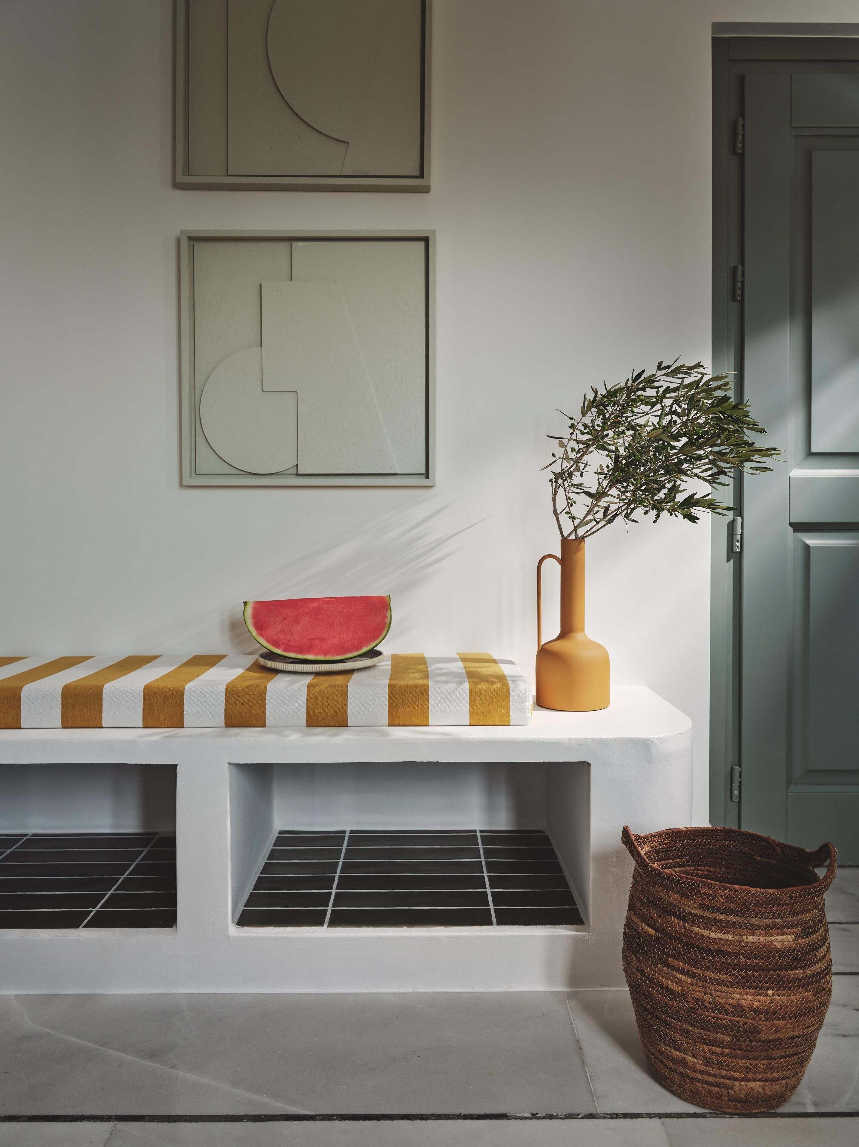 A white bench with a striped cushion, a vase with a plant, and a woven basket.