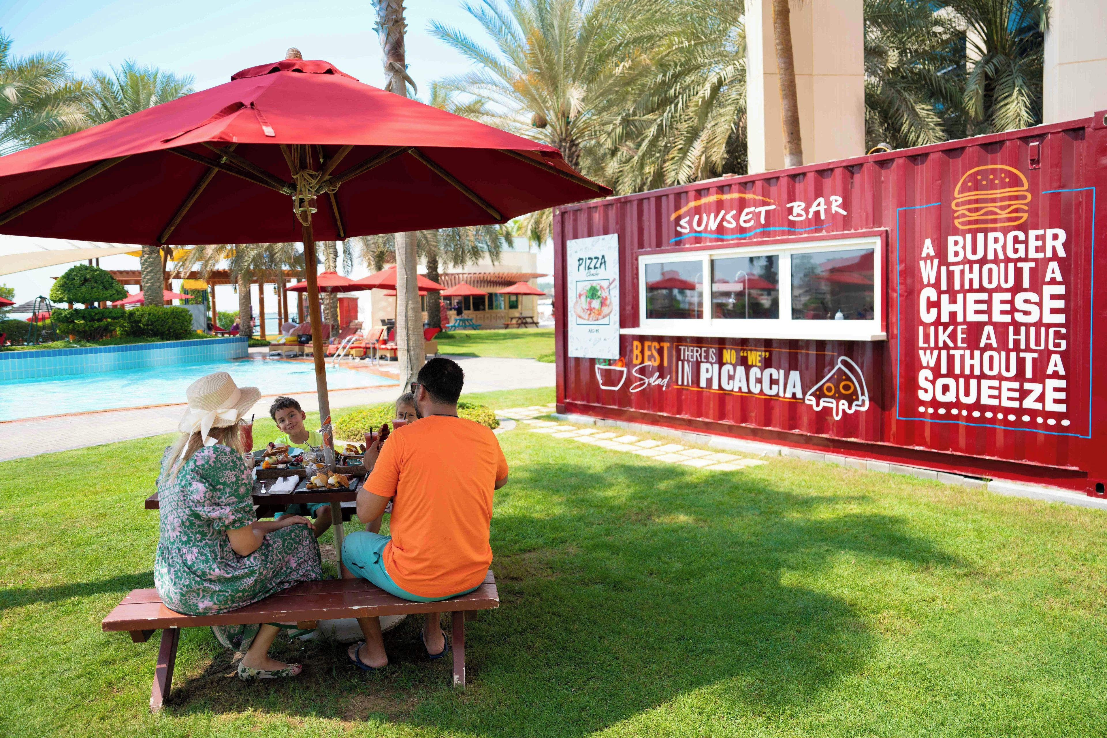 A group of people dining at a restaurant with a red umbrella, a pool in the background, and a sign advertising a burger without cheese.