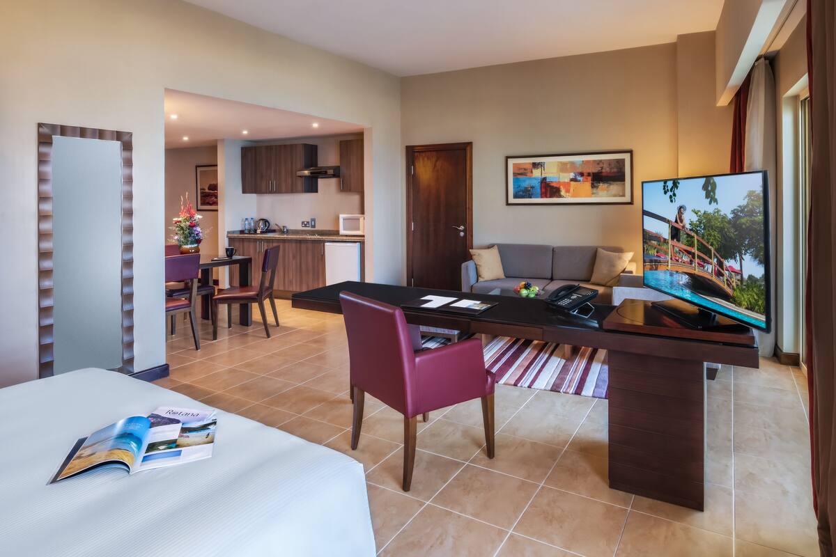 A hotel room with a dining area, a kitchen, a living area with a TV, and a door leading to another room.