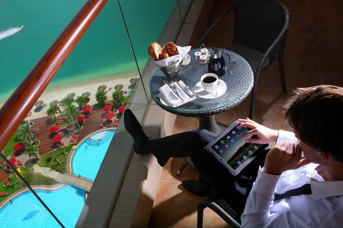 A man sitting at a table with a tablet, a cup of coffee, and a basket of pastries. The table is on a balcony with a view of a pool and a beach.
