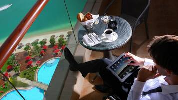 A man sitting at a table with a tablet, a cup of coffee, and a basket of pastries. The table is on a balcony with a view of a pool and a beach.