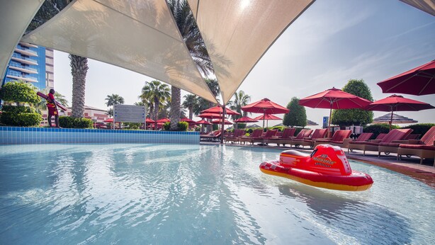 A swimming pool with red umbrellas and lounge chairs, surrounded by palm trees and a blue building. - Khalidiya Palace Rayhaan by Rotana (Abu Dhabi)