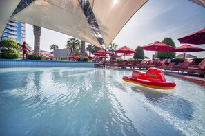 Una piscina con sombrillas rojas y sillas de descanso, rodeada de palmeras y un edificio azul.