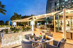 Un área de comedor al aire libre con mesas y sillas, luces colgantes y una vista de un edificio al fondo.