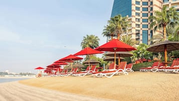 Une plage avec des parasols rouges et des chaises longues blanches, un grand immeuble en arrière-plan et des palmiers.