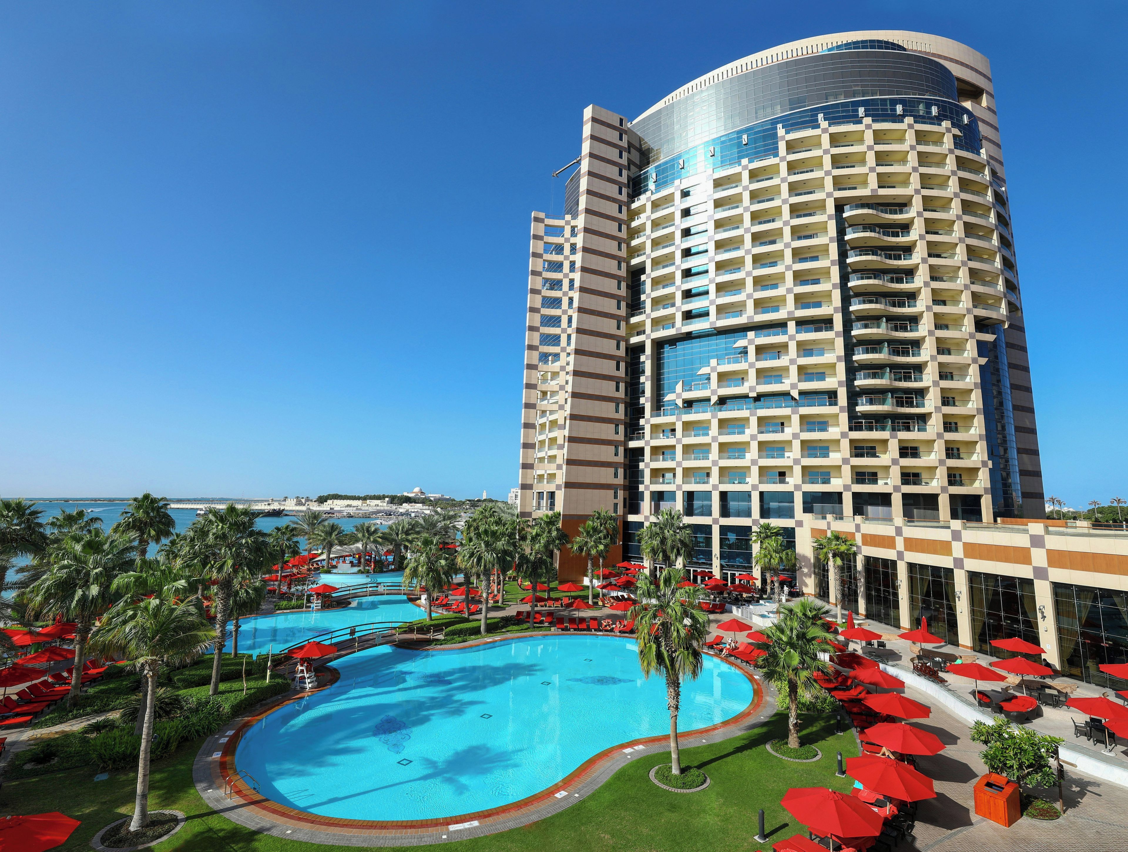 Un hôtel doté d’une grande piscine, de palmiers et de parasols rouges.
