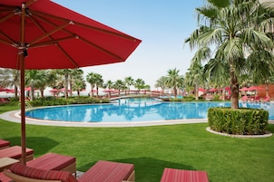 A swimming pool surrounded by palm trees and red umbrellas, with lounge chairs and a wooden bridge.
