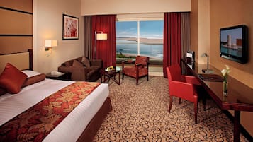 A hotel room with a bed, a desk, a chair, a TV, and a window with a view of the beach.