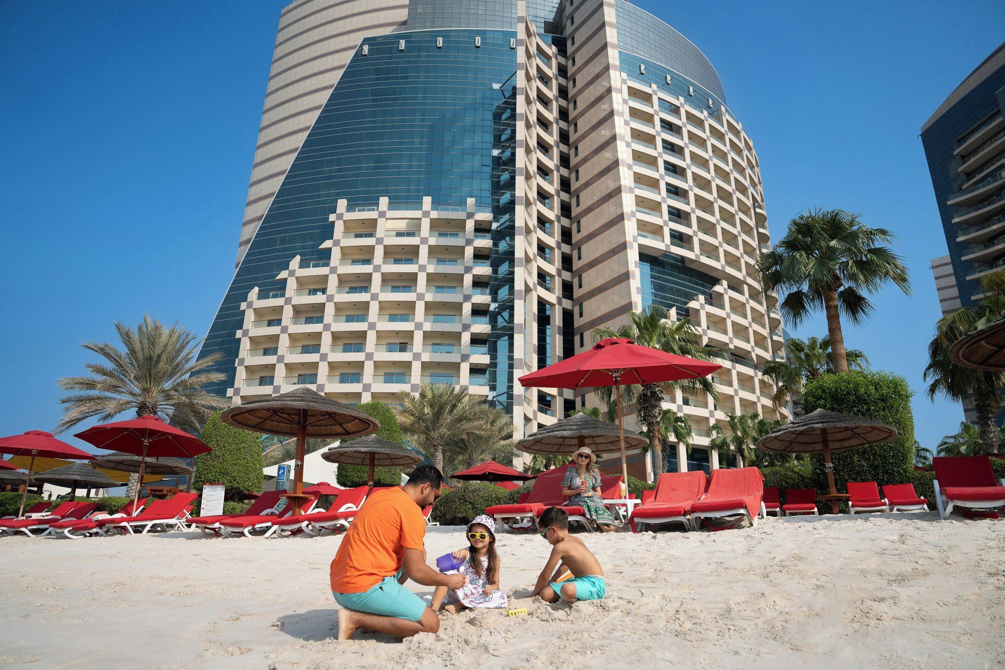 Une plage avec des parasols rouges, des chaises longues et un bâtiment moderne en arrière-plan.