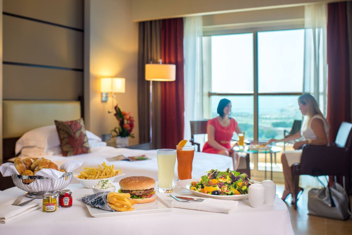 A hotel room with a dining table set with food, a bed, and a view of the city.