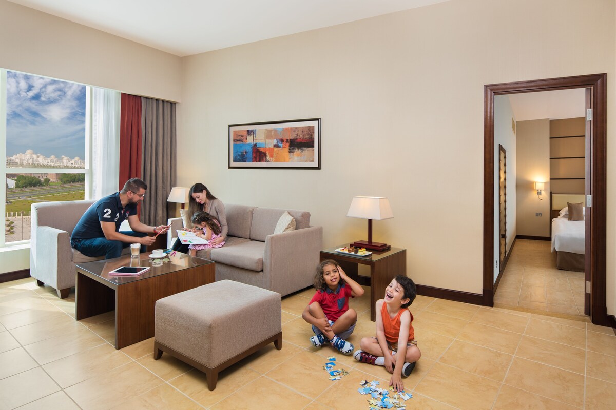 A hotel room with a large window, a sofa, a coffee table, and a child playing with puzzle pieces.