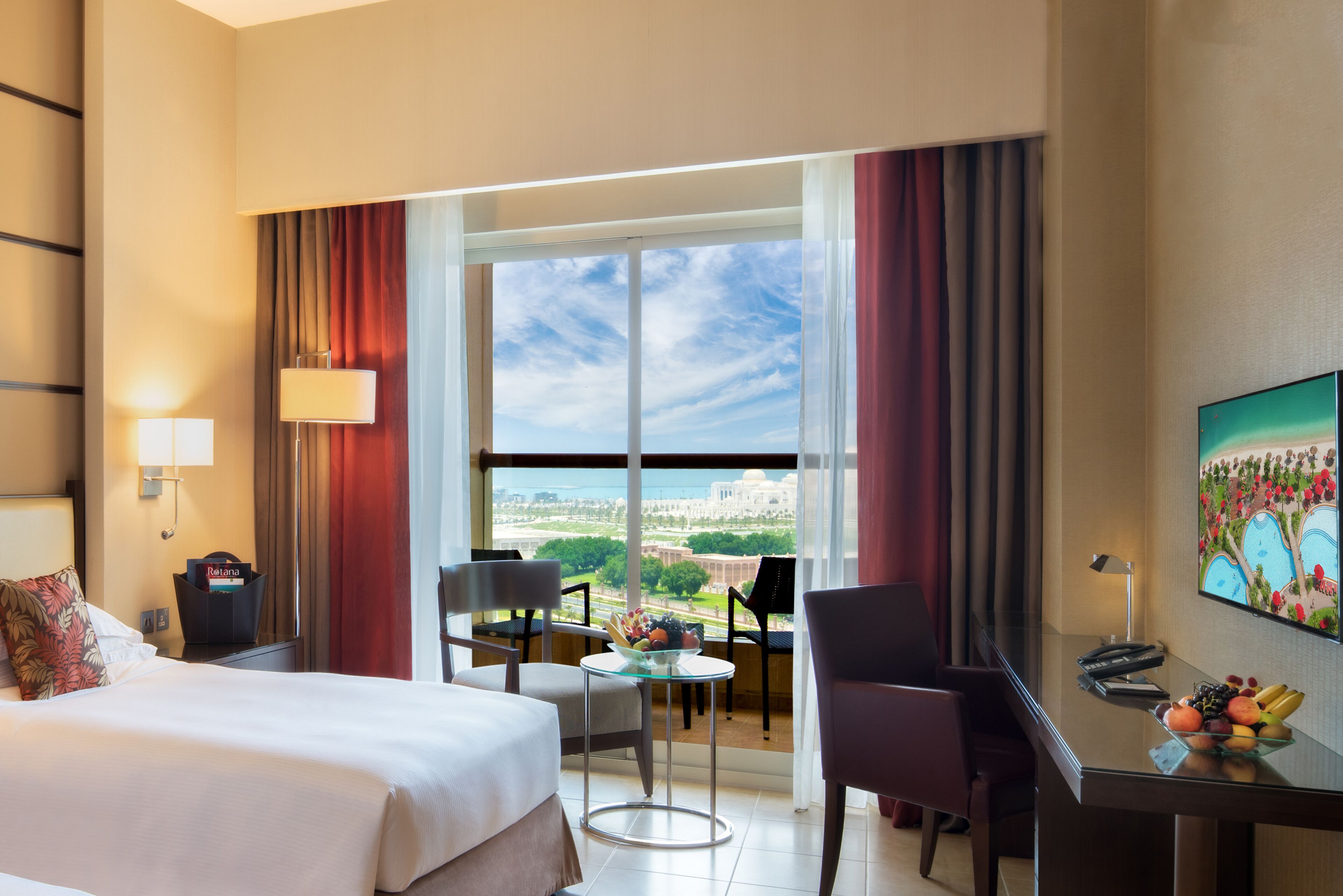 A hotel room with a bed, a desk, a chair, a small table, and a large window offering a view of a cityscape.