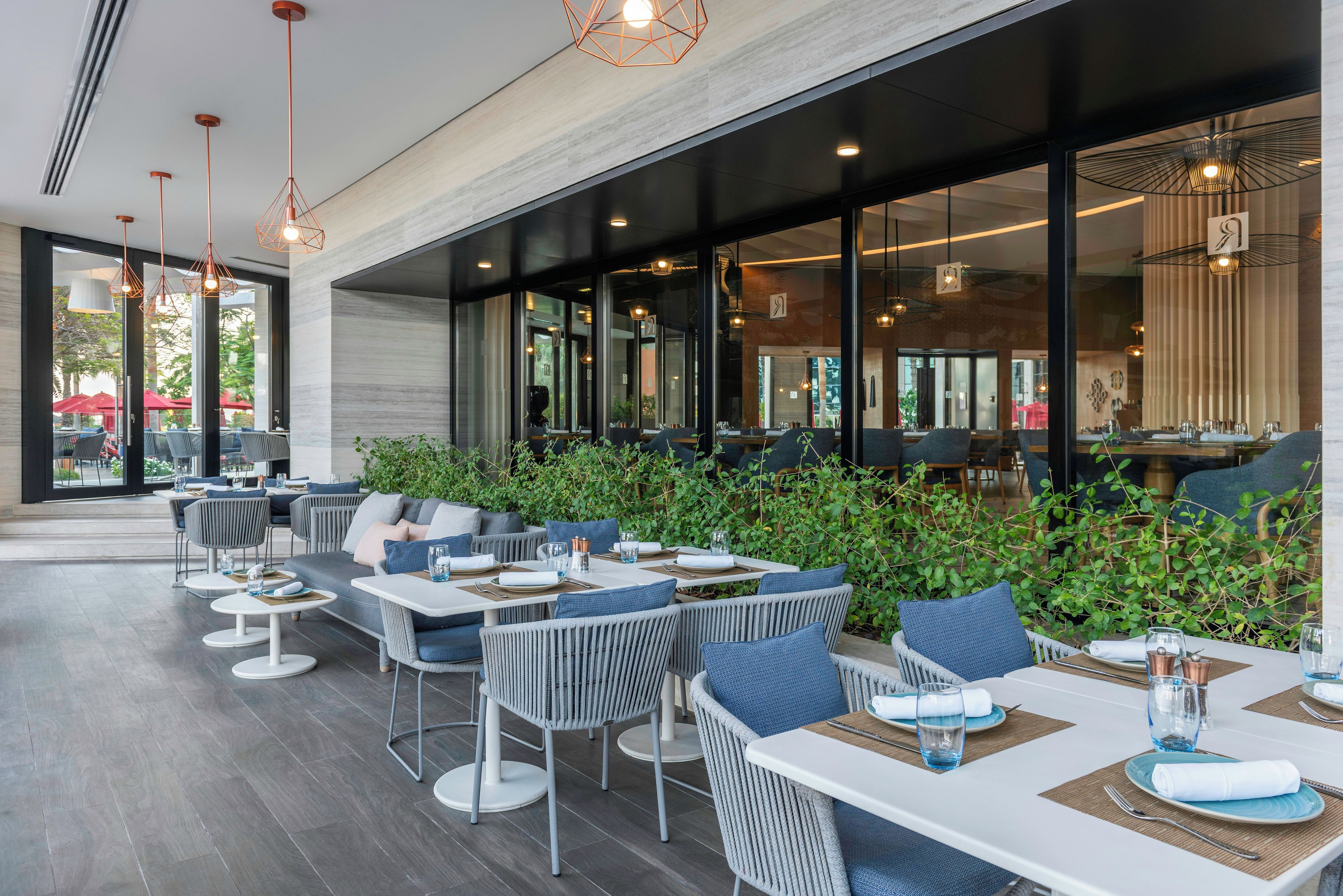 An outdoor dining area with modern furniture, large windows, and greenery.