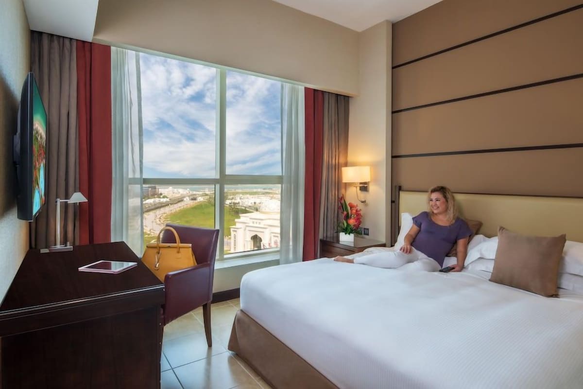 A hotel room with a large window, a bed, a desk, and a chair.