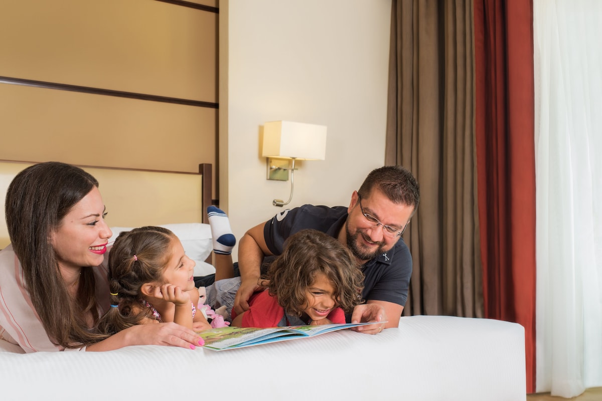 A family of four lying on a bed in a hotel room, reading a book together.