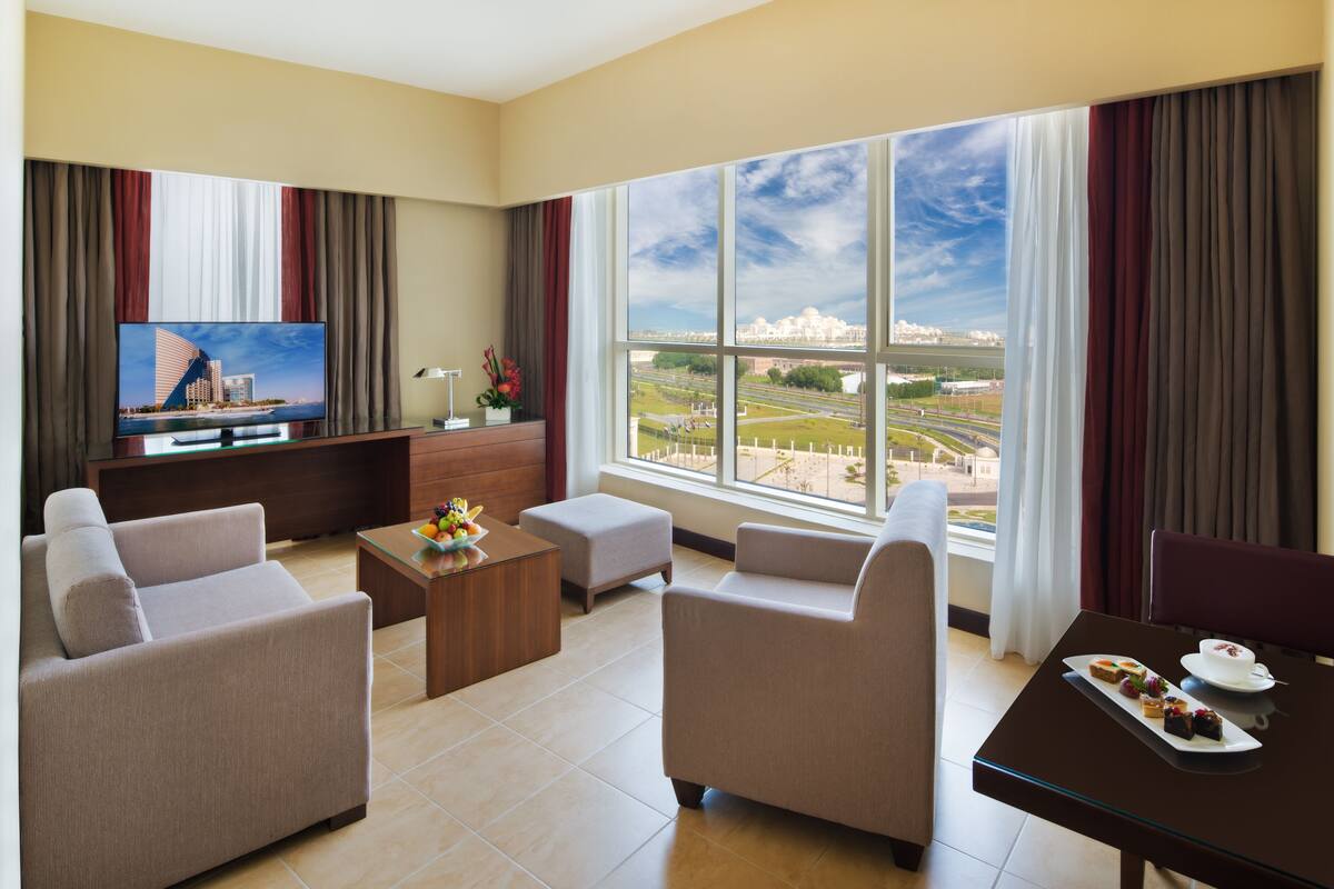 A modern hotel room with a large window offering a city view, a flat-screen TV, and a dining area with a table set for two.
