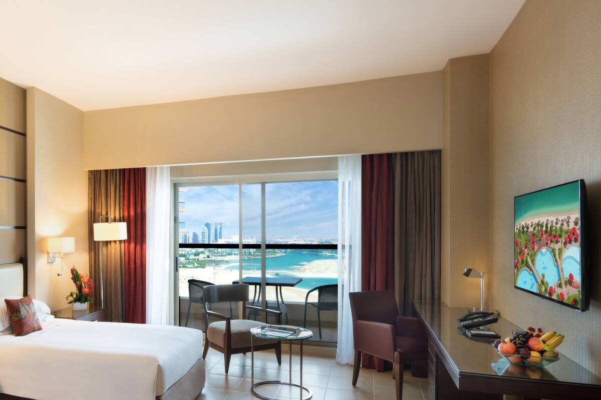 A hotel room with a bed, a desk with a fruit bowl, a chair, a table, and a view of a beach and cityscape through large windows.